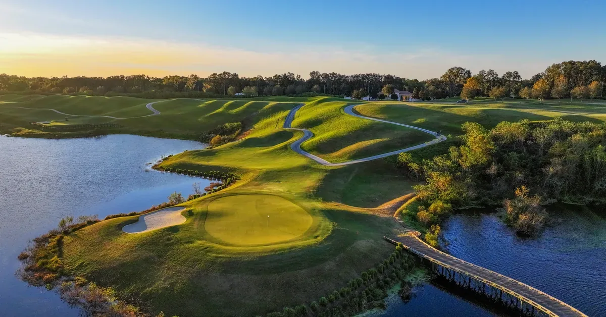 Aerial sunset view of a lush golf course featuring rolling fairways, sand bunkers, and a scenic wooden bridge spanning a tranquil water hazard.